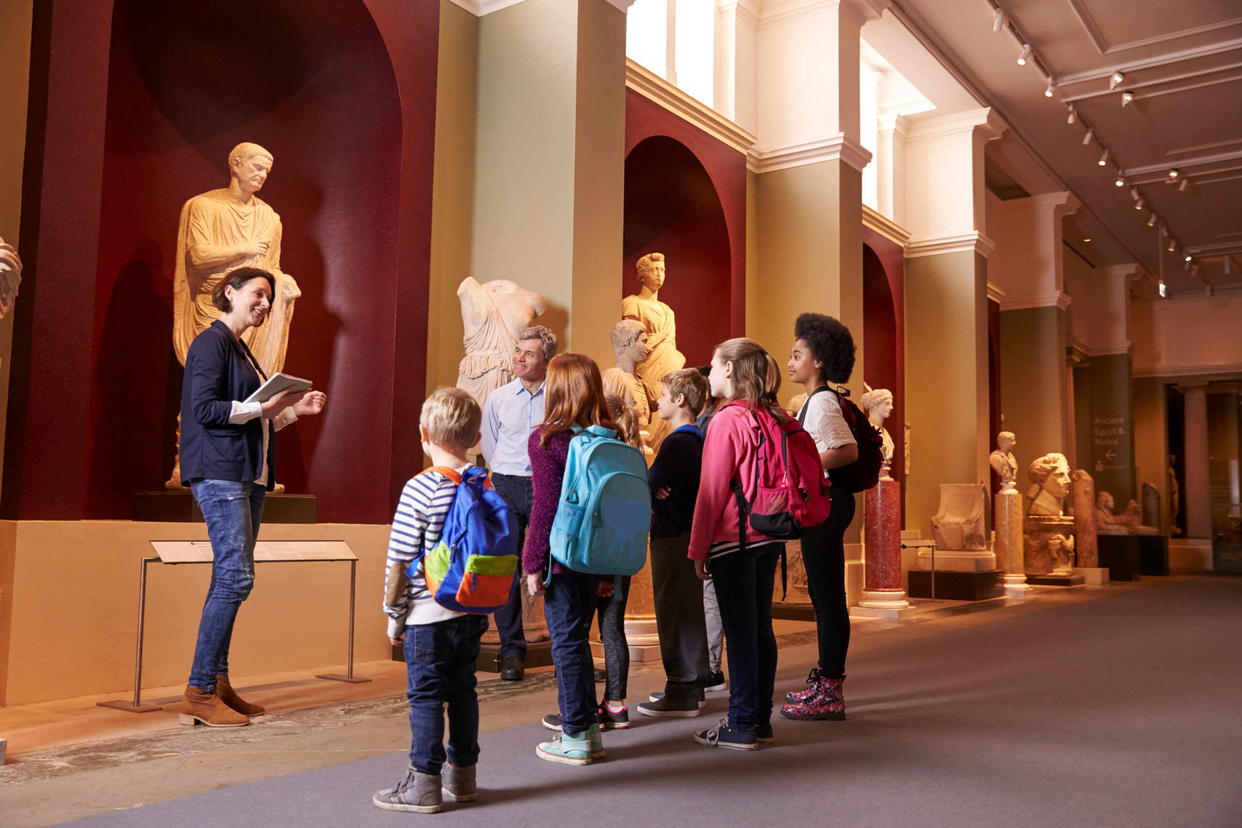Gruppe von Schulkindern hört einer Museumsführerin bei antiken Skulpturen aufmerksam zu.