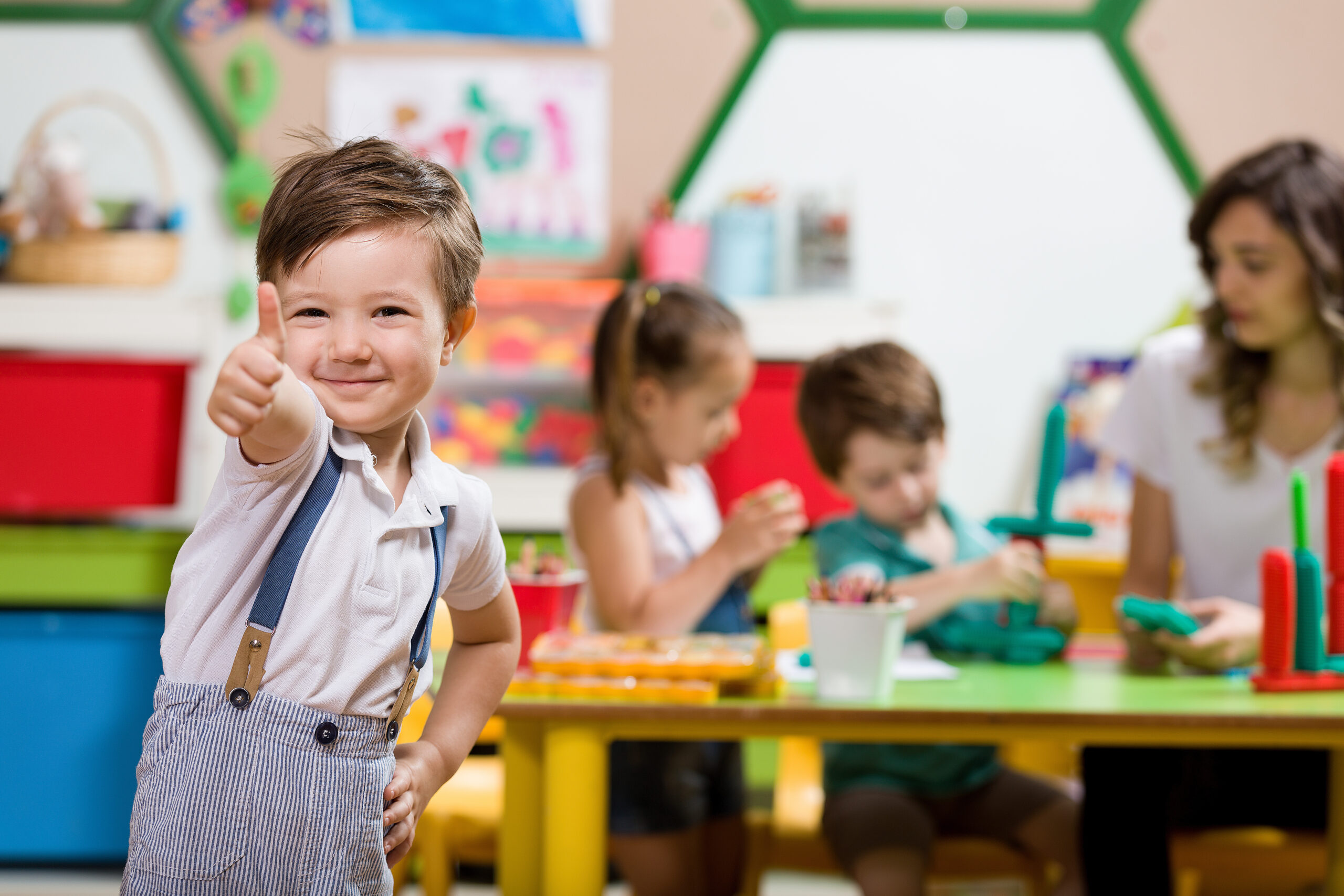 Lächelnder Junge zeigt Daumen hoch, während Kinder im Kindergarten gemeinsam spielen.