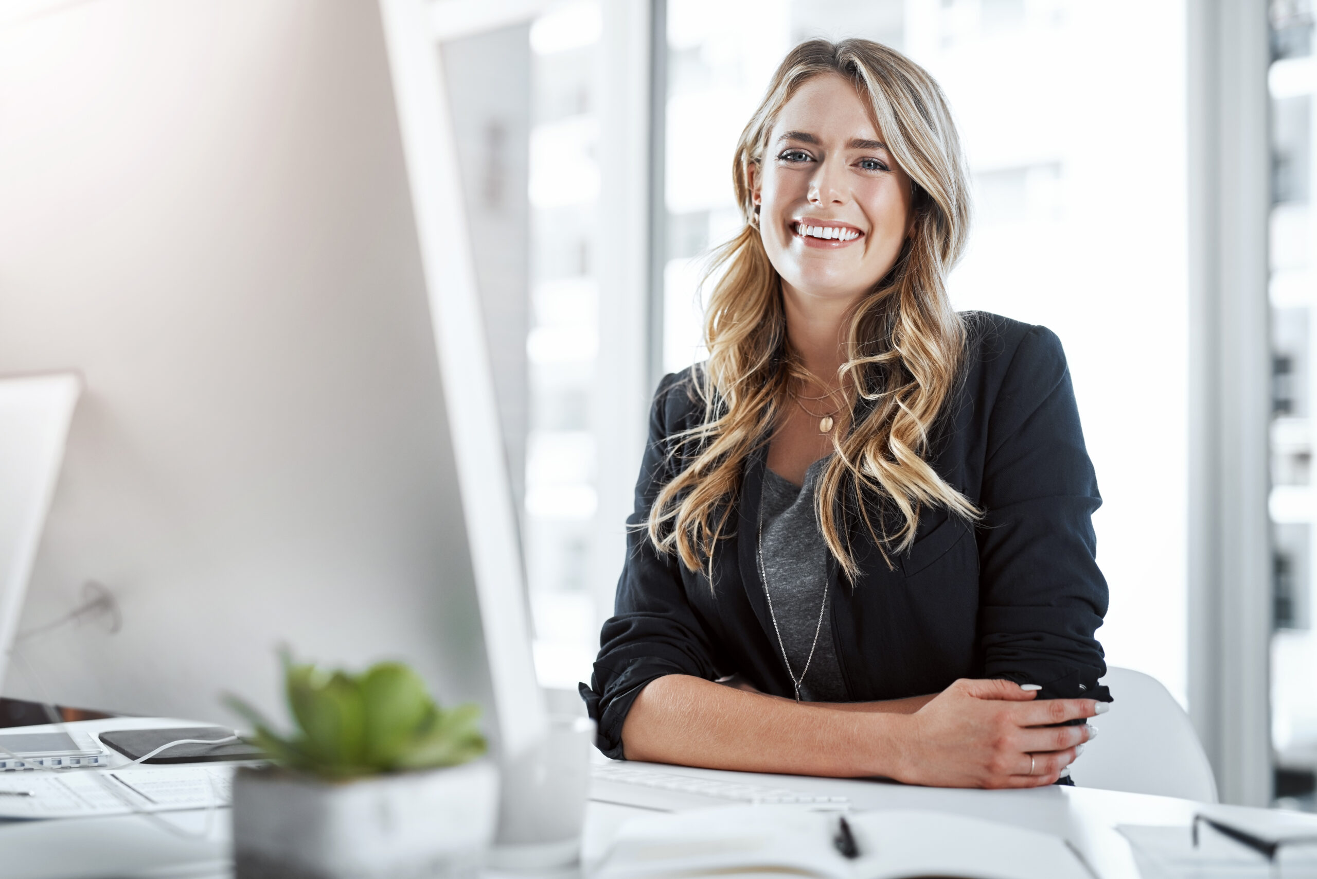 Fachkraft für Finanzen sitzt im Business-Outfit lächelnd am Schreibtisch vor Computer im hellen Büro.