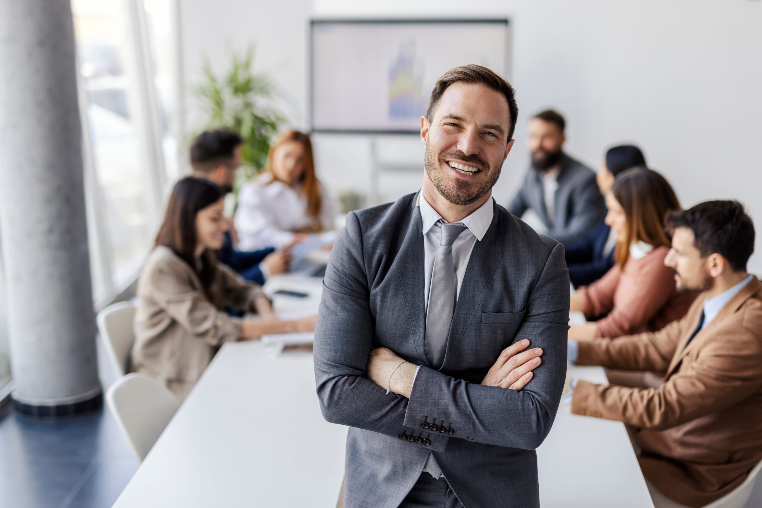 Eine Person steht mit verschränkten Armen im Vordergrund eines Besprechungsraums, während im Hintergrund ein Team gemeinsam an einem Tisch arbeitet und Unterlagen bespricht.